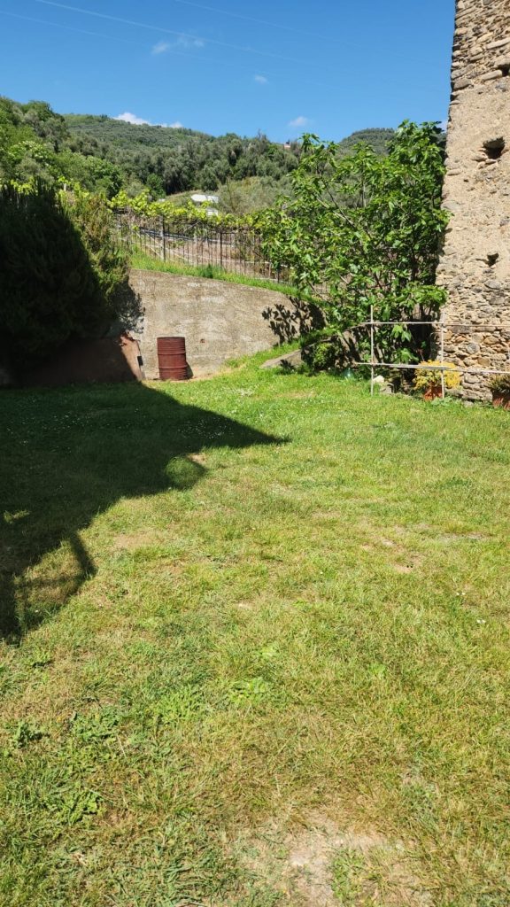 Giardino pianeggiante tra casa in pietra e muro di contenimento, con vista sulle colline circostanti.